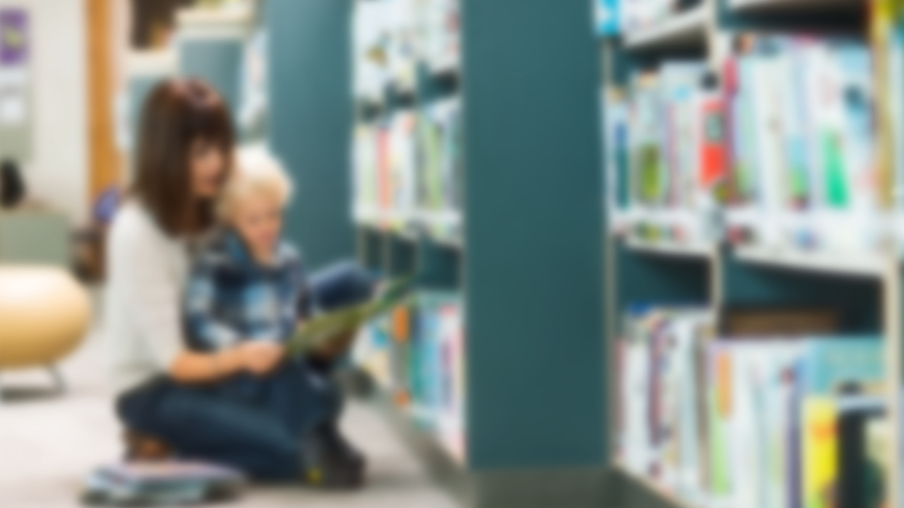 Blurred adult sitting on library floor reading to a child who is sitting on her lap. There are library shelves with books to the right.