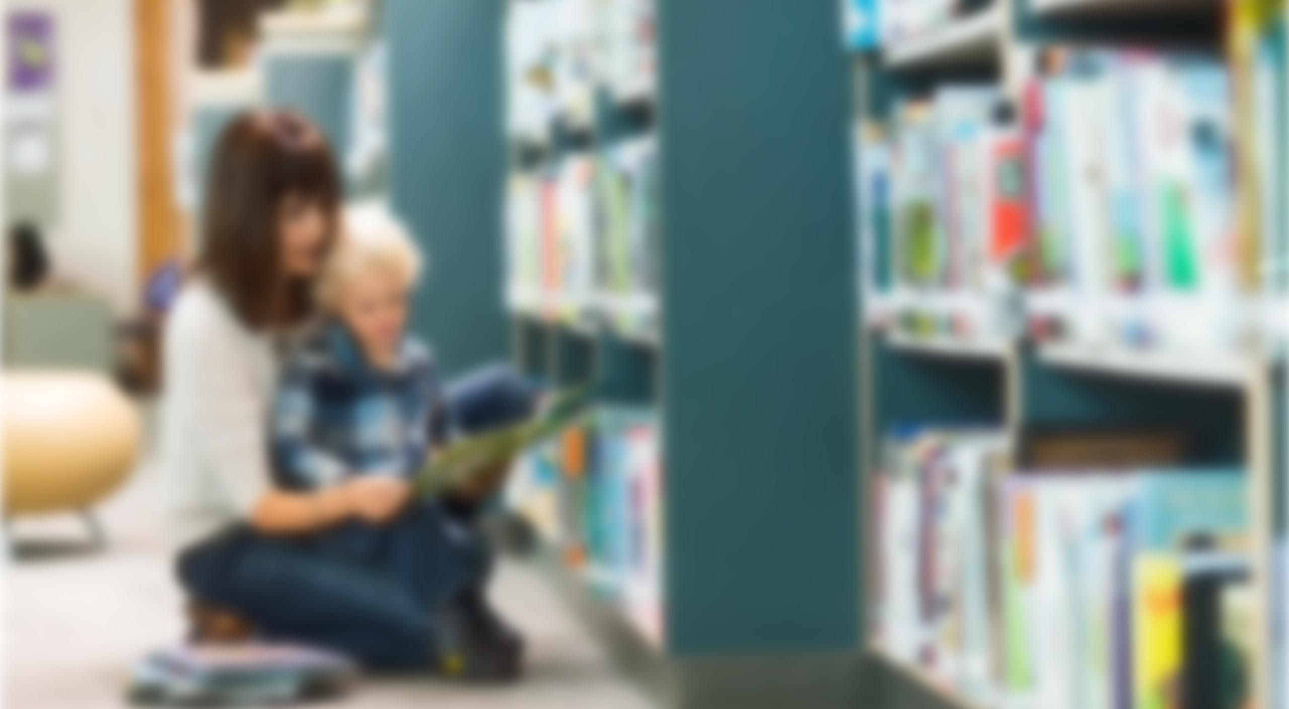 Blurred adult sitting on library floor reading to a child who is sitting on her lap. There are library shelves with books to the right.
