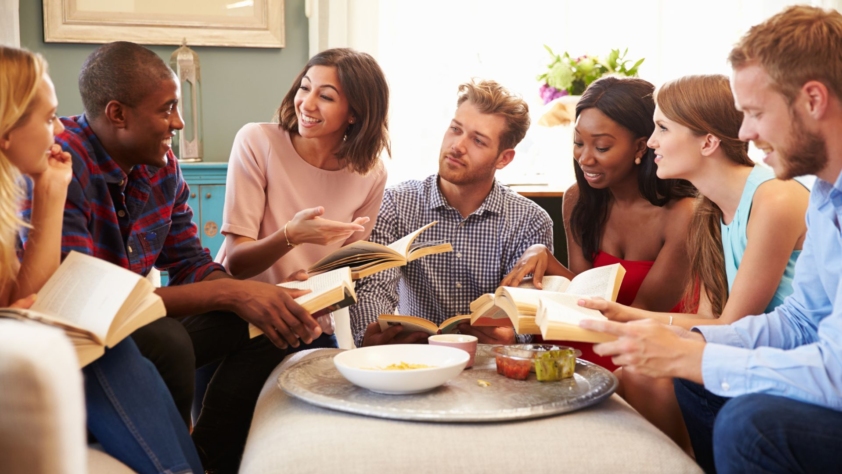 Book Clubs and Writing Groups events photo. Seven people are sitting around a large ottoman holding books and talking. There is food on the ottoman.