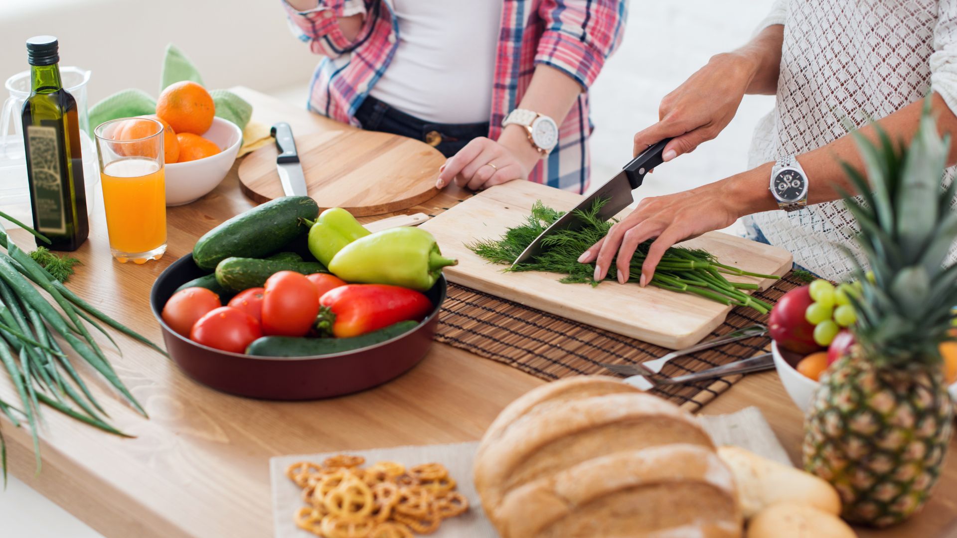 Health and Wellbeing events photo. Two people are standing at a counter and are preparing food. One person is cutting herbs on a butting board. There are oranges in a bowl, juice in a class, a bottle of oil, as well as other fruit, vegetables, pretzels and a loaf of bread.