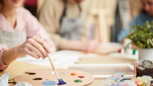 Hobbies, Crafts and Games events photo. A person is painting at a table. There are two more people at the table but they are farther away from the camera and are blurry.