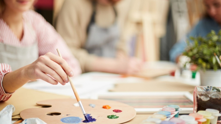 Hobbies, Crafts and Games events photo. A person is painting at a table. There are two more people at the table but they are farther away from the camera and are blurry.