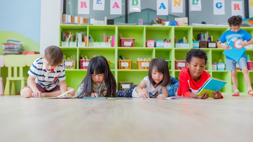 Kids and Families events photo. There are 5 children in the photo. They are either in a library or classroom. All children are viewing books. One child is sitting on the floor, three children are lying on the floor, and one child is standing. There are shelves behind them with baskets of books.