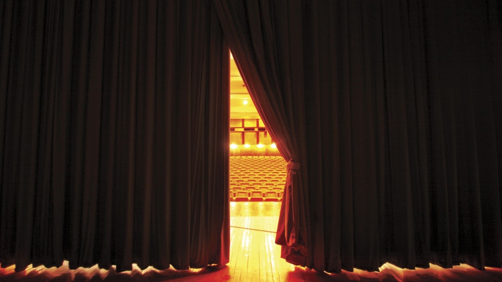 Special events photo. The picture is from the perspective of someone behind the curtains on a stage. There is a small opening in the curtains and the theatre seats are visible.