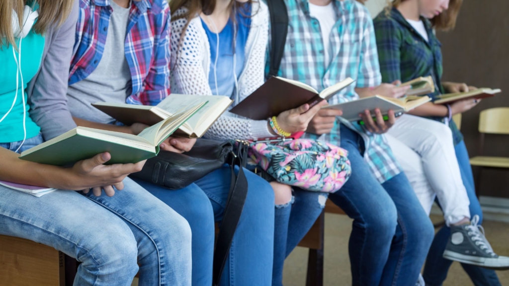 Teens events photo. Five teenagers are sitting on a table or desk reading bools, while one teenager stands at the end of the table, also reading a book.