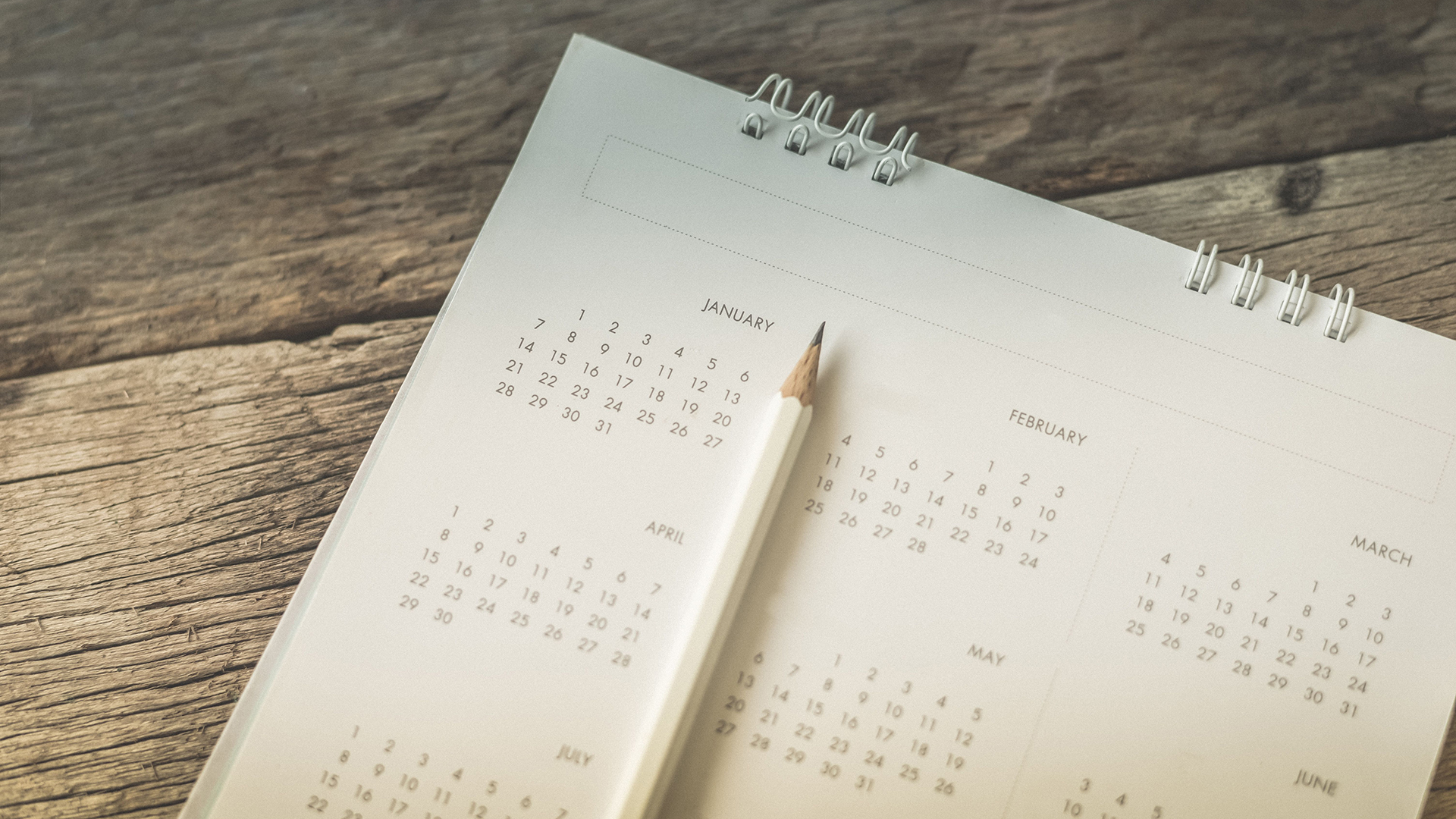 Holiday and Seasonal events image. There is a white calendar containing some of the months of a year with a white pencil laying on top of it. The calendar is placed on a wood background.