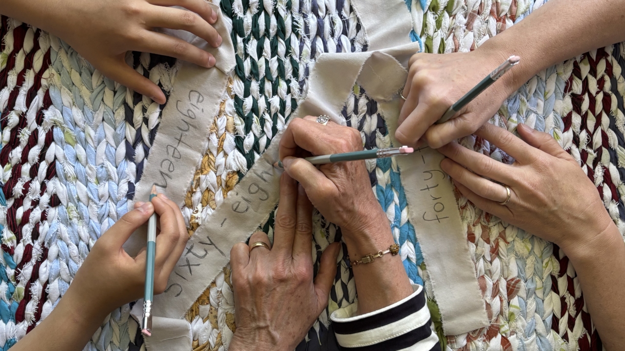 Photo image shows 4 pairs of hands writing on strips of fabric on top of a weaving.