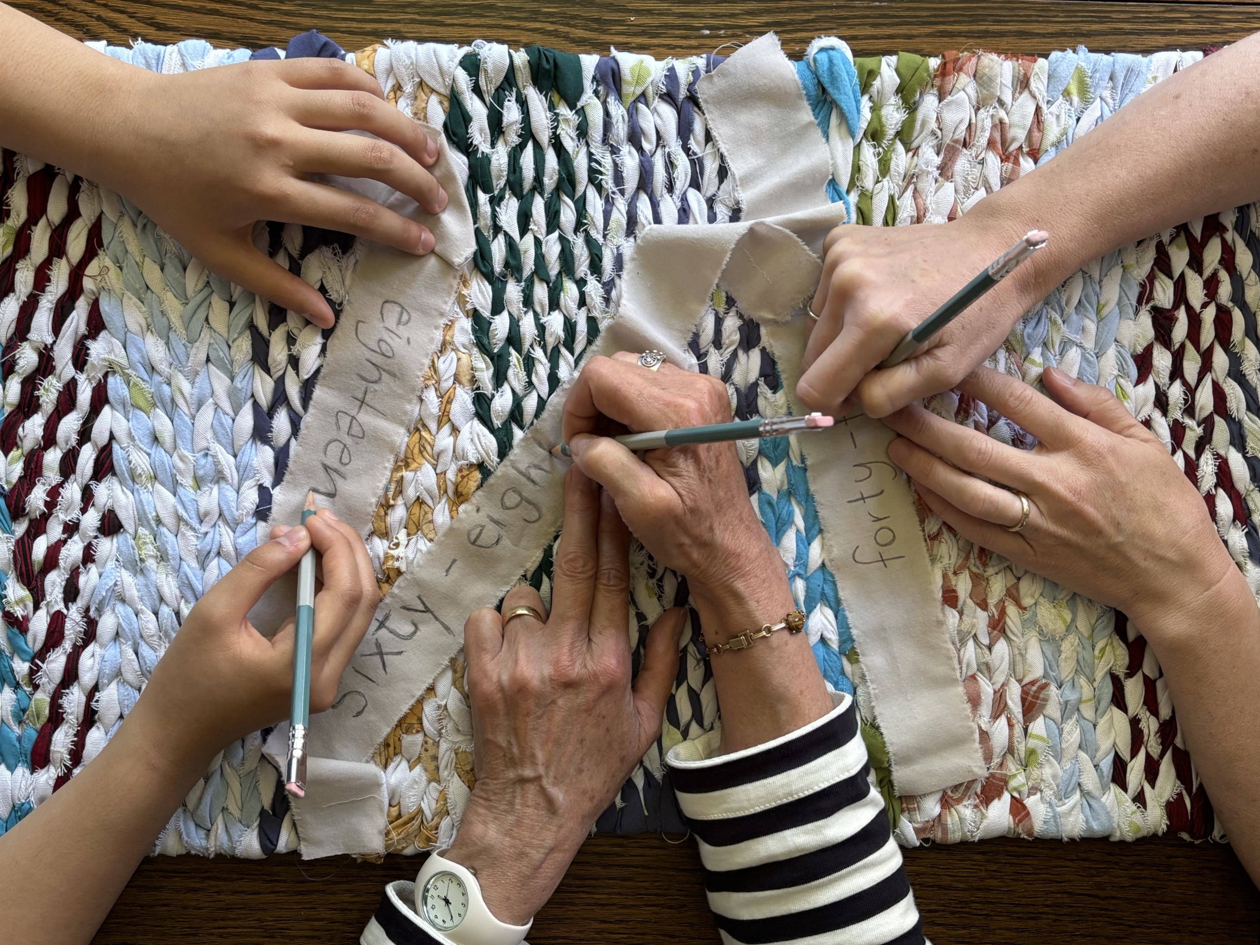 Photo image shows 4 pairs of hands writing on strips of fabric on top of a weaving.